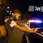 Młody mężczyzna, który został zatrzymany do kontroli policyjnej, opiera się dłońmi o swój samochód. Policjant go spisuje. Jest noc.